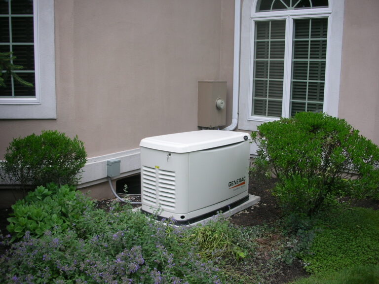 A standby generator is installed outside a house, positioned near bushes and a window with white trim, showcasing a professional home generator installation.