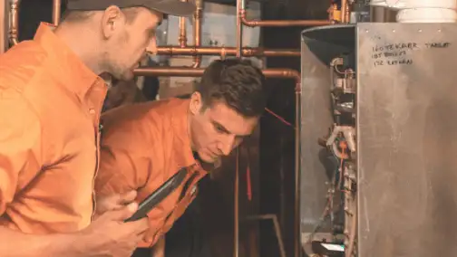 Two technicians in orange shirts conduct a general heating repair, inspecting a furnace closely. One technician holds a tool while copper pipes gleam subtly in the background.
