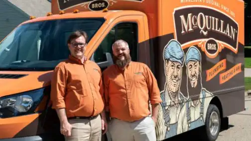 Two men in orange shirts stand proudly in front of a McQuillan Bros van, showcasing graphics of two illustrated workers, ready to tackle any general heating repair needs with expertise and efficiency.