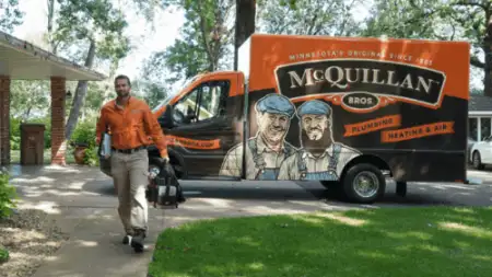 A technician in an orange shirt walks from a McQuillan Bros service van, which is parked on a driveway. The van, showcasing plumbing and heating services branding, specializes in general heating repair to ensure your home stays comfortable year-round.