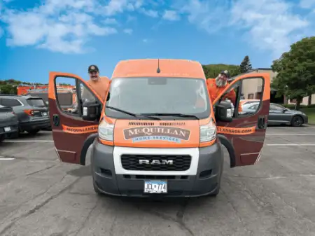 Two people stand by an orange McQuillan Home Services van with open front doors in a parking lot, ready for their next general heating repair task.