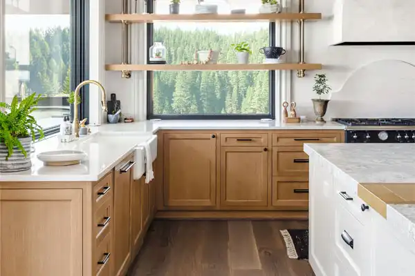 Modern kitchen with light wood cabinets, white countertops, and open shelving adorned with potted plants. A large window offers a view of green trees, while expertly designed plumbing ensures smooth operations without the need for repair.