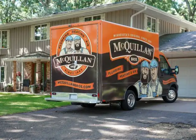 An orange McQuillan Bros plumbing truck, specializing in General Heating Repair, is parked in the driveway of a beige house, framed by lush trees in the background.