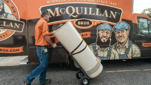 A worker expertly transports a large water heater on a dolly, passing by a McQuillan Bros van adorned with branding and illustrations, showcasing their expertise in general heating repair.