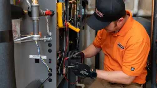 A technician wearing a cap and gloves focuses on general heating repair, expertly adjusting wires and tools inside the furnace.