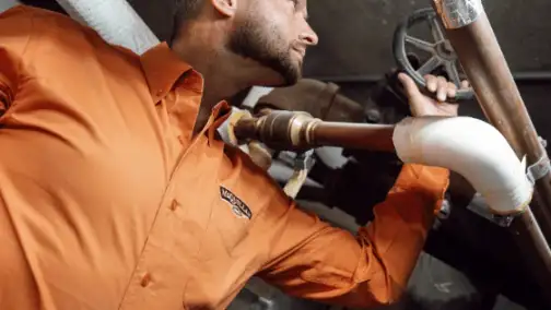 A man in an orange shirt is inspecting industrial pipes and turning a valve, focusing on general heating repair duties.
