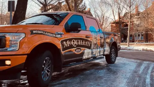 The orange McQuillan Bros truck, known for its General Heating Repair expertise, is parked on a snowy street with trees and houses in the background.