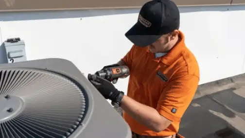 A technician in an orange shirt and black hat skillfully uses a power tool to perform general heating repair on an outdoor air conditioning unit.