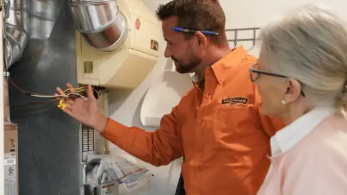 A technician in an orange shirt is demonstrating HVAC wiring to a woman with gray hair, as they both inspect the furnace unit for general heating repair in a utility room.
