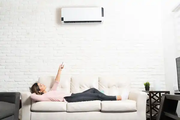 A person lounging on a sofa effortlessly operates a wall-mounted air conditioner installed through an efficient mini-split installation, set against a backdrop of white brick walls.