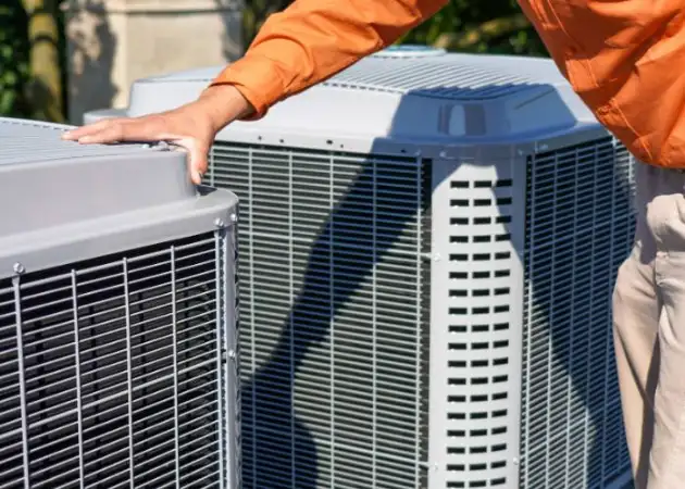 A person in an orange shirt carefully inspects two outdoor air conditioning units placed side by side, ensuring everything is in top condition for general heating repair.